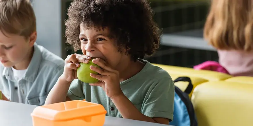 Young child taking a big bite of a green apple at a cafeteria table, with an orange lunch box in front and a baby blue backpack behind. Two other children are visible in the background.