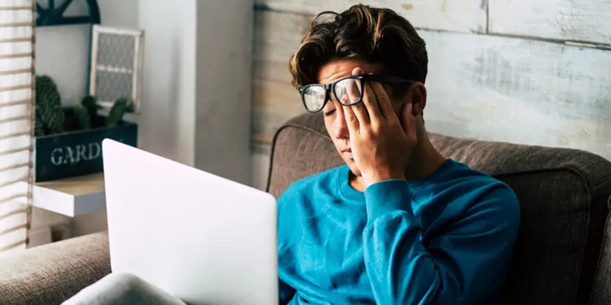 Someone in glasses sitting on a couch looking tired and unwell while using a laptop, showing possible COVID symptoms like fatigue or headache.