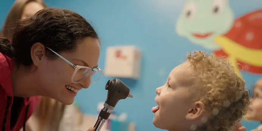 Smiling healthcare provider examining a young child’s throat with an otoscope during a friendly urgent care visit.